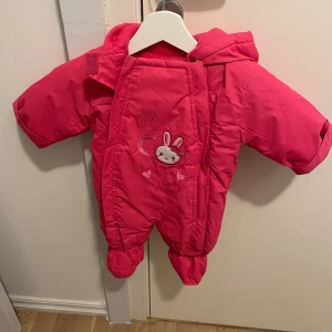 Rosa overall med kaninmotiv - Söt rosa overall med kaninmotiv och hjärtdetaljer. Perfekt för att hålla de små varma under kyliga dagar. Overallen har en praktisk dragkedja och huva för extra skydd mot väder och vind.