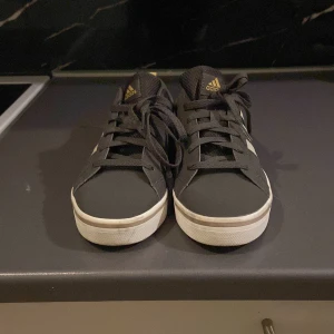 Svarta sneakers från Adidas - Snygga svarta sneakers från Adidas med vita ränder och detaljer. Skorna har en klassisk design med snörning och en vit sula. Perfekta för en avslappnad stil.