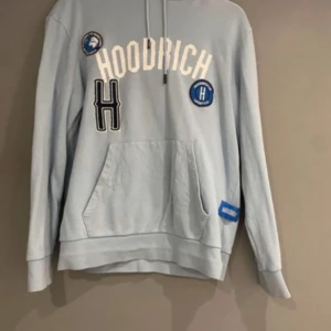 Ljusblå hoodie från Hoodrich - Säljer en ljusblå hoodie från Hoodrich med tryck och patchar på framsidan. Tröjan har en stor ficka framtill och justerbar huva med dragsko. Perfekt för en avslappnad stil.