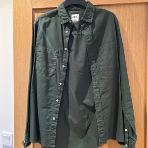 Grön långärmad skjorta från ZARA - Snygg grön långärmad skjorta från ZARA med klassisk krage och knappar framtill. Skjortan har två bröstfickor och en avslappnad passform, perfekt för en stilren look.
