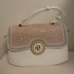 Vit handväska med glittrig detalj - Säljer en elegant vit handväska med glittrig framsida och gulddetaljer. Väskan har ett dekorativt spänne och ett praktiskt handtag. Insidan är rymlig med dragkedja för säker förvaring. Perfekt för att ge en touch av glamour till din outfit!