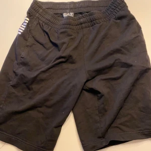 Svarta shorts från Emporio Armani storlek S - Snygga svarta shorts från Emporio Armani EA7 med elastisk midja och randiga detaljer på sidan. Perfekta för en avslappnad stil med en touch av lyx. Märkeslogga synlig på benet.