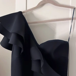 Svart fest/bal klänning - Elegant svart klänning med en stilren design och volangdetaljer. Klänningen är väldigt stretchig och har en asymmetrisk axel och en smickrande passform som ger en sofistikerad look. Perfekt för speciella tillfällen, användes endast en gång.