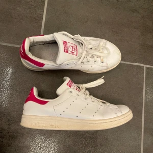 Adidas Stan Smith sneakers i vitt och rosa - Säljer ett par klassiska Adidas Stan Smith sneakers i vitt med rosa detaljer. Skorna har snörning och den ikoniska Stan Smith-loggan på plösen och hälen