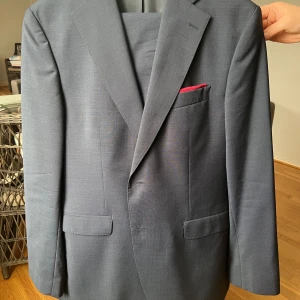 Mörkblå kavaj från Sir of Sweden - Elegant mörkblå kostym set från Sir of Sweden med klassisk passform. Kavajen har två knappar framtill och en stilren bröstficka med en röd näsduk. Perfekt för formella tillfällen.