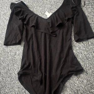Svart offshoulder body med volanger - Säljer en svart offshoulder body från H&M med volangdetaljer. Toppen har trekvartsärm och en elegant design som passar perfekt för olika tillfällen.