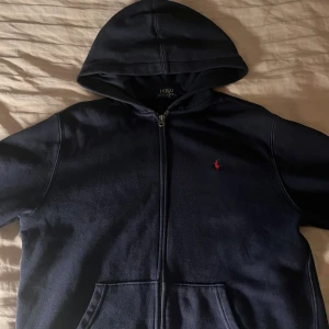 Mörkblå hoodie från Polo Ralph Lauren - Säljer en snygg mörkblå hoodie från Polo Ralph Lauren med dragkedja och huva. Den har den ikoniska röda loggan på bröstet. Passar både S och M.