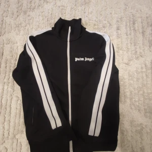 Palm Angels Kofta - Säljer en svart track jacket från Palm Angels med vita ränder längs ärmarna och dragkedja framtill. Jackan har en hög krage och Palm Angels-logga på bröstet. Perfekt för en sportig look!