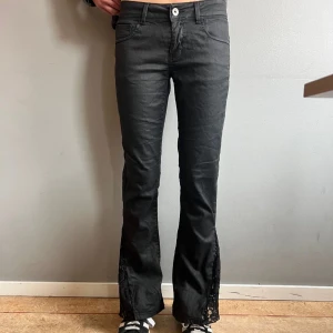 Svarta bootcut byxor med spetsdetaljer - Snygga svarta bootcut byxor med unika spetsdetaljer längs benen. Byxorna har en knapp och dragkedja framtill och är perfekta för en trendig look. De är lågmidjade och har en cool stil som sticker ut. Storlek: small, midjemått 38cm, innerbenslängd 76cm💕