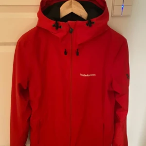 Röd vindjacka från Peak Performance - Snygg röd skidjacka från Peak Performance med dragkedja och justerbar huva. Perfekt för blåsiga dagar och utomhusaktiviteter. Jackan har en stilren design med diskret logga på bröstet.