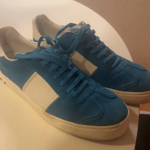 Valentino Flycrew Blåa - Snygga blå och vita sneakers från Valentino med coola nitar på hälen. Skorna har en klassisk snörning och är tillverkade i mocka och skinn. Perfekta för att ge din outfit en extra edge.