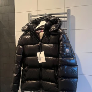 Moncler Maya  - Säljer en snygg svart pufferjacka från Moncler. Jackan har en glansig finish och är utrustad med en dragkedja framtill. Den har en praktisk huva och Monclers logotyp på ärmen. Perfekt för kyliga dagar! Finns både nfc Scan samt qr kod, Jackan har en liten repa som knappast syns om du inte kollar noga men därav det låga priset+icke säsong spara in nu och köp en billigt tills nästa vinter säsong. Självklart äkta men inget äkhetskapsbevis.