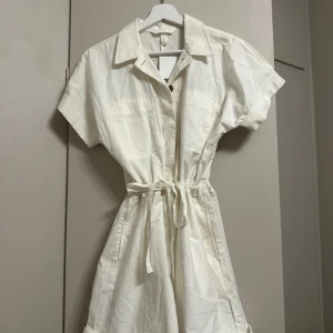 Vit kortärmad playsuit med knytband - Säljer en stilren vit playsuit med korta ärmar och knytband i midjan. Den har en klassisk krage och knappar framtill. Perfekt för en avslappnad sommarlook.