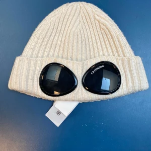 Vit ribbad mössa från C.P. Company - Snygg vit ribbad mössa från C.P. Company med unika glasögondetaljer framtill. Perfekt för att hålla stilen under kyliga dagar. Mössan har en modern design och är tillverkad i ett mjukt material för extra komfort.