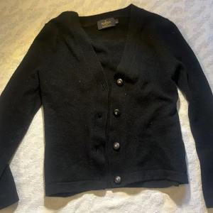 Svart kofta från Morris Lady - Säljer en elegant svart kofta från Morris Lady i 100% merinoull. Koftan har en v-ringad design med knappar framtill och långa ärmar. Perfekt för att hålla dig varm och stilren under kyligare dagar.