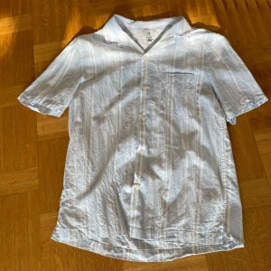 Vit och ljusblå randig skjorta från H&M - Säljer en stilren vit och ljusblå randig skjorta från H&M. Skjortan har korta ärmar och knappar framtill. Perfekt för en avslappnad look. 🕶️