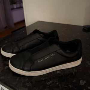 Svarta sneakers från Tommy Hilfiger - Snygga svarta sneakers från Tommy Hilfiger med en stilren design. Skorna har en vit sula och är tillverkade i skinn. Perfekta för en casual look. Dom är använda med nåra få skrapor. men det går att tvätta bort vilket jag gör innan dom skickas! 