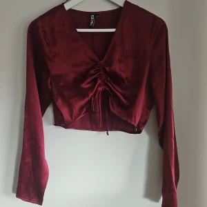 Vinröd satinblus från Pieces - Snygg vinröd blus i satin från Pieces med långa ärmar och en v-ringad design. Blusen har en trendig snörning framtill som ger en fin detalj och är croppad för en modern look.