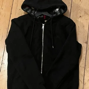 Svart hoodie från Burberry - Säljer en svart hoodie från Burberry med dragkedja och klassiskt rutigt mönster på insidan av huvan. Perfekt för en casual look med en touch av lyx. Den har långa ärmar och en bekväm passform.