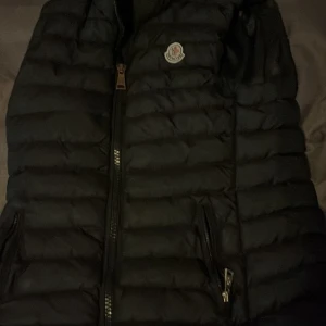 Svart dunväst från Moncler - Snygg svart dunväst från Moncler med dragkedja och logotyp på bröstet. Perfekt för kyligare dagar och ger en stilren look. Västen har en quiltad design och praktiska fickor med dragkedja. Storleken passar M och S