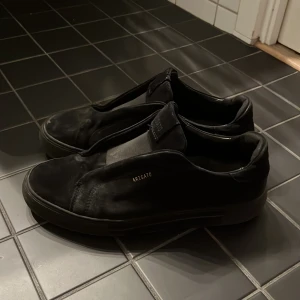 Svarta sneakers från Axel Arigato - Snygga svarta sneakers från Axel Arigato i skinn. De har en stilren design utan snörning och en diskret logga på sidan. Perfekta för en minimalistisk look. Lite sliten sula men annars i bra skick