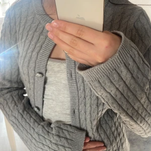 Grå stickad kofta med knappar - Säljer en mysig grå stickad kofta med kabelstickat mönster och dekorativa knappar. Perfekt för kyliga dagar och ger en stilren look. Passar bra över en enkel topp eller klänning. Passar på folk som har storlek S också. 💓💓