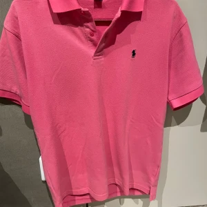 Rosa pikétröja från Ralph Lauren - Säljer en snygg rosa pikétröja från Ralph Lauren i storlek L. Tröjan är tillverkad i 100% bomull och har en klassisk krage med knappar. Perfekt för en stilren look!