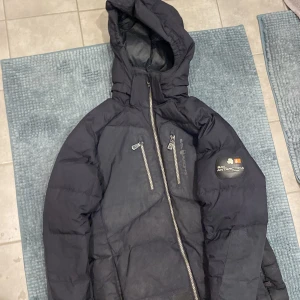 Svart dunjacka från Sail Racing - Säljer en svart dunjacka från Sail Racing med huva och dragkedja. Jackan har en logotyp på bröstet och är designad för kalla klimat. Perfekt för vinterns äventyr och håller dig varm och bekväm.