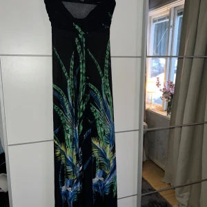 Svart axelbandslös klänning med mönster från Stella - Snygg svart axelbandslös långklänning från Stella med ett tropiskt mönster i grönt, blått och gult. Perfekt för en somrig look med sin flowiga design och färgglada detaljer. 🌿