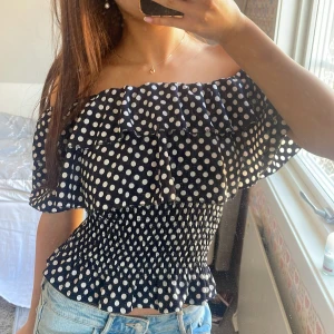 Svart offshoulder topp med prickar - Säljer en söt svart offshoulder topp med vita prickar. Toppen har volangdetaljer och en smockad midja som ger en fin passform. Perfekt för en somrig look! 