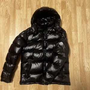 Moncler Maya - Säljer en snygg svart dunjacka från Moncler med huva och dragkedja. Jackan har ett stilrent Moncler-märke på ärmen och är perfekt för kyliga dagar. Den är både varm och trendig, idealisk för vintern. Det är en 1, 1 Maya i väldigt bra skick den är i nyskick i storlek medium
