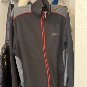 Svart jacka från Boss - Säljer en stilren svart jacka från Boss med grå och röda detaljer. Jackan har en dragkedja framtill och långa ärmar. Perfekt för en casual look.