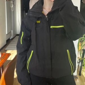 Svart jacka från Helly Hansen - Säljer en svart jacka från Helly Hansen med neongula dragkedjor och detaljer. Jackan har en normal passform och är perfekt för utomhusaktiviteter. Den har flera praktiska fickor med dragkedjor och en hög krage för extra skydd. Dragkedja är lite seg.