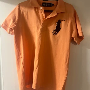 Orange pikétröja från Ralph Lauren - Säljer en snygg orange pikétröja från Ralph Lauren med den klassiska loggan på bröstet. Tröjan har korta ärmar och en krage med knappar. Perfekt för en stilren och avslappnad look.