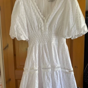 By Malina klänning - Supersöt vit sommarklänning med puffärmar, v-ringning och broderade spetsdetaljer. Klänningen har ett diskret prickigt mönster. Perfekt för sommarens alla tillfällen!