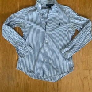 Randig skjorta från Polo Ralph Lauren - Snygg blå- och vitrandig skjorta från Polo Ralph Lauren med klassisk krage och broderad logga på bröstet. Custom fit-modell med långa ärmar och knappar framtill. Perfekt för dig som gillar stilrena och tidlösa plagg.