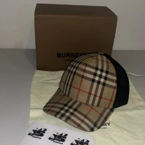 Burberry rutig keps med mesh - Snygg keps från Burberry med klassiskt rutigt mönster i beige, svart och rött framtill och svart mesh baktill. Justerbar snapback och böjd skärm. Perfekt accessoar för en trendig streetwear-look.