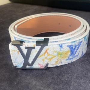 Vit Louis Vuitton bälte med färgglatt mönster - Säljer ett vitt Louis Vuitton bälte i skinn med färgglada LV-loggor och monogram i blått, gult, rött och grönt. Bältet har en stor silverfärgad LV-spänne och exklusiv känsla. Perfekt för dig som vill sticka ut med en lyxig accessoar.