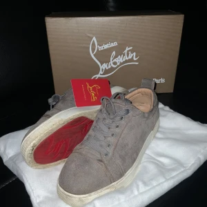 Grå mocka sneakers från Louboutin - Snygga grå sneakers från Christian Louboutin i mjuk mocka med klassisk röd sula. Skorna har rund tå, grå snörning och vita sulor. Insidan är beige och de har en stilren, minimalistisk look som passar till många outfits. Skorna är i väldigt bra skick men finns dock några defekter. Som ni ser på bild 4 så är det röd färgprick som jag inte vet hur man får bort, sen så är det typ en liten öppning på bild 5 i skon. Men inget som störs. Allt OG medföljer. Osäker om äktheten och skorna passar 39-40.