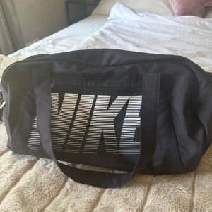 Svart träningsväska från Nike - Säljer en svart träningsväska från Nike med stort vitt logotryck på sidan. Väskan har rejäla handtag och är tillverkad i slitstark polyester. Perfekt för gymmet eller sportaktiviteter, rymmer allt du behöver och är enkel att bära med sig.
