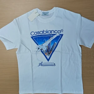 Vit Casablanca t-shirt med tryck - Vit t-shirt från Casablanca med blått grafiskt tryck framtill som visar ett flygplan och stadssilhuett. Klassisk rund hals och korta ärmar. Skön och mjuk i bomull, perfekt för en clean och modern look.