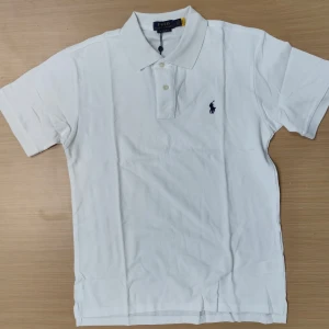 Vit pikétröja från Polo Ralph Lauren - Klassisk vit pikétröja från Polo Ralph Lauren med korta ärmar och broderad marinblå logga på bröstet. Tröjan har krage och två knappar framtill. Tillverkad i bomull för en skön och luftig känsla. Perfekt för en clean och stilren look. Vid minsta fundering skicka dm :)