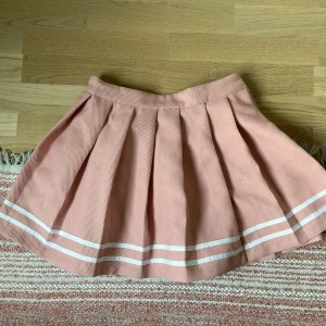 Rosa plisserad kjol - Superfin rosa plisserad kortkjol från H&M Divided med vita ränder längst ner. Kjolen har dragkedja i sidan och knapp i midjan. Köpte den i en blindbox för 3 år sedan men den passade inte så har bar legat i min garderob och anser att den är alldeles för fin för detta. Tjockt och stadigt tyg