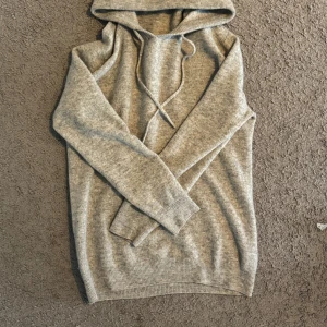 Grå stickad hoodie med snörning - Mysig grå hoodie i stickat material med huva och snörning framtill. Tröjan har långa ärmar och ribbade muddar vid ärmslut och nederkant. Perfekt för kyliga dagar och enkel att matcha med jeans eller mjukisbyxor. Men har tyvärr att hål under armen men syns inte alls 