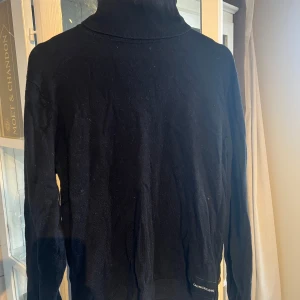 Svart polotröja Calvin Klein Jeans - Svart polotröja från Calvin Klein Jeans i kashmirblandning. Klassisk design med ribbade muddar och hög krage. Diskret logga på nedre delen av tröjan. Perfekt för dig som gillar stilrena och bekväma plagg.