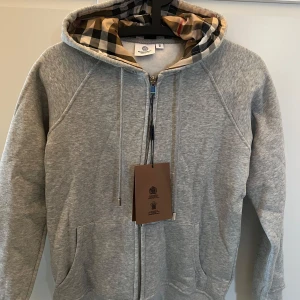 Grå hoodie Burberry - Snygg grå hoodie från Burberry i bomull med klassiskt rutigt foder i huvan. Tröjan har dragkedja framtill, snörning vid huvan och två fickor. Perfekt för dig som vill ha en stilren och lyxig vardagslook. Sitter som en S/M 