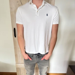 Vit pikétröja från Ralph Lauren - Klassisk vit pikétröja från Ralph Lauren med korta ärmar och den ikoniska broderade loggan på bröstet. Tröjan har krage, knappar framtill och är gjord i mjuk bomull. Perfekt för en clean och stilren look.