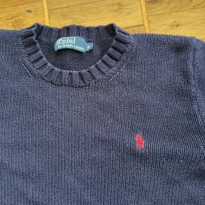 Mörkblå stickad tröja Polo Ralph Lauren - Mörkblå stickad tröja från Polo Ralph Lauren med klassisk rund hals och ribbade muddar. Tröjan har det ikoniska röda Polo-logot broderat på bröstet. Perfekt för dig som gillar stilren och tidlös design.