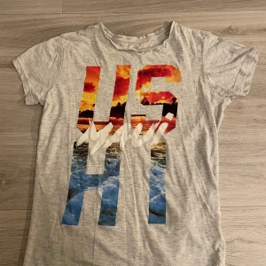 Grå  t-shirt med tryck - Snygg grå t-shirt i ekologisk bomull från Cubus med stort färgglatt tryck på framsidan där det står 'US HI Maui' med strand- och solnedgångsmotiv. Klassisk passform och korta ärmar, perfekt för en chill sommarlook.