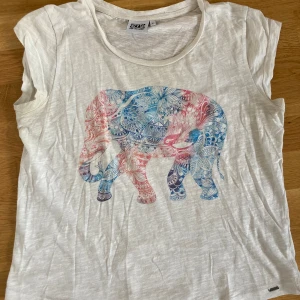 Vit t-shirt med elefanttryck från Animal - Vit t-shirt från Animal med ett stort, detaljerat elefanttryck i blått och rosa på framsidan. T-shirten är ärmlös med rund halsringning och tillverkad i tunn, mjuk bomull. Perfekt för dig som gillar unika prints och chill stil.